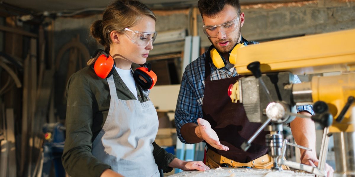 Waist-up portrait of attractive apprentice standing at drill press and listening to her male mentor with attention, he explaining her operating principle of machine