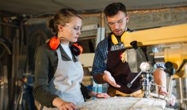 Waist-up portrait of attractive apprentice standing at drill press and listening to her male mentor with attention, he explaining her operating principle of machine